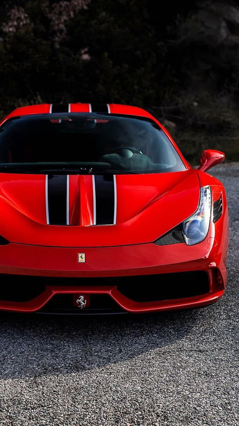 Red Ferrari sports car with black and white racing stripes parked on a gravel road in a frontal portrait view.