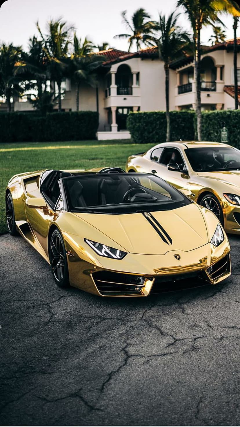 Gold Lamborghini convertible and coupe parked before a luxury villa with palm trees under bright sunlight.