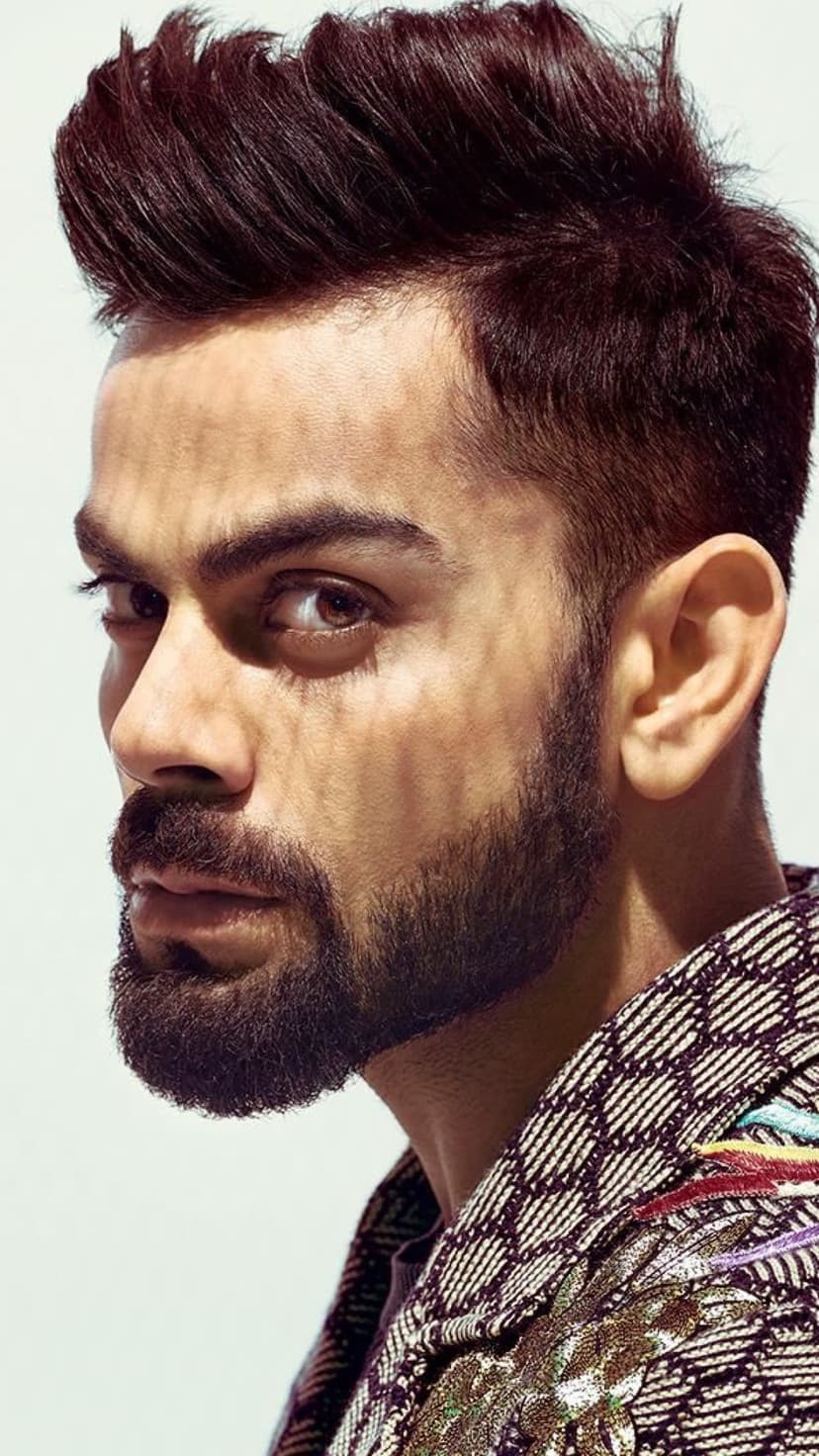 Virat Kohli in a side profile view wearing a patterned jacket with a groomed beard and dramatic side lighting.