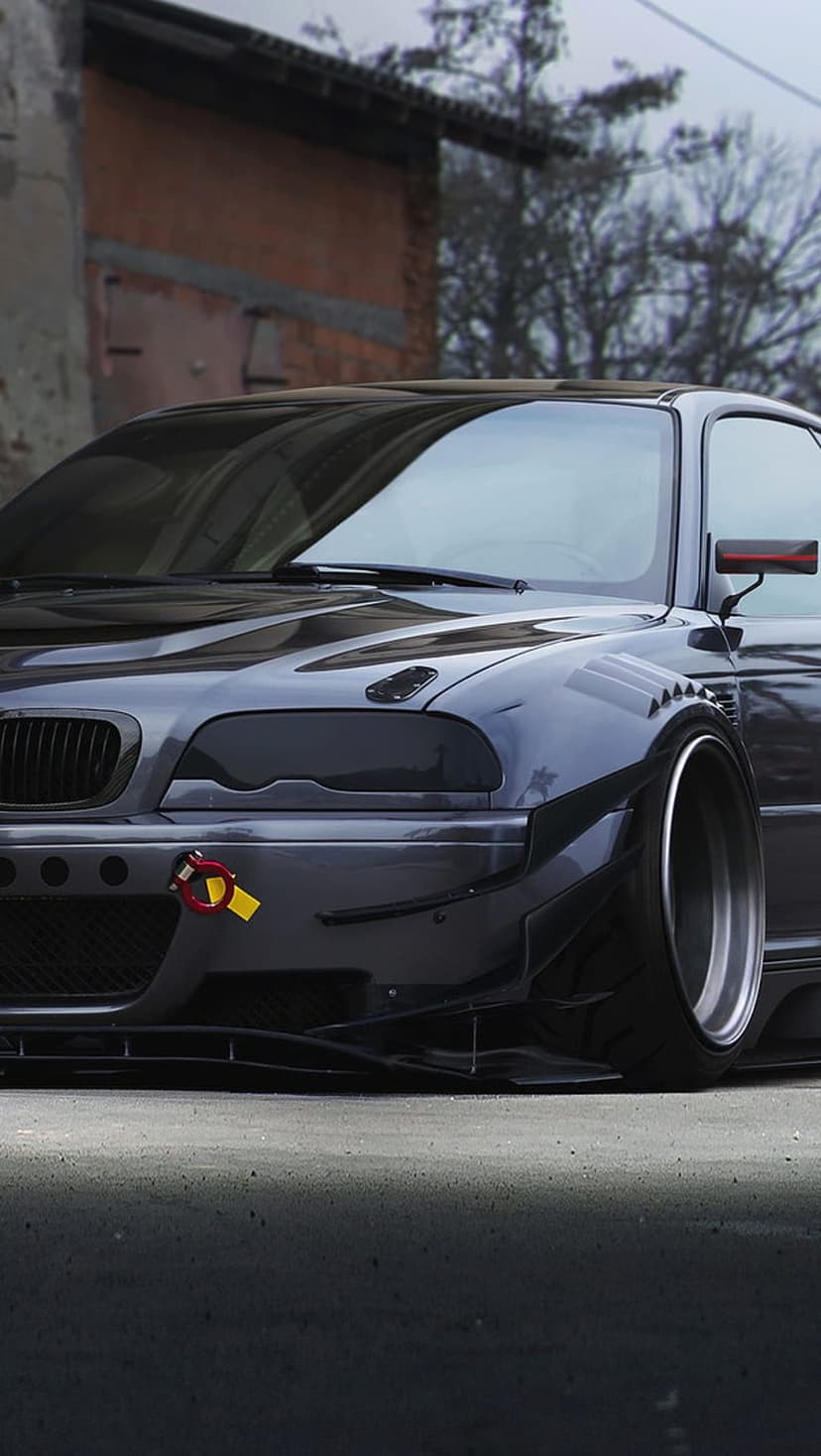 BMW E46 M3 with flared widebody fenders and blacked-out headlights features a lowered stance and gunmetal paint.