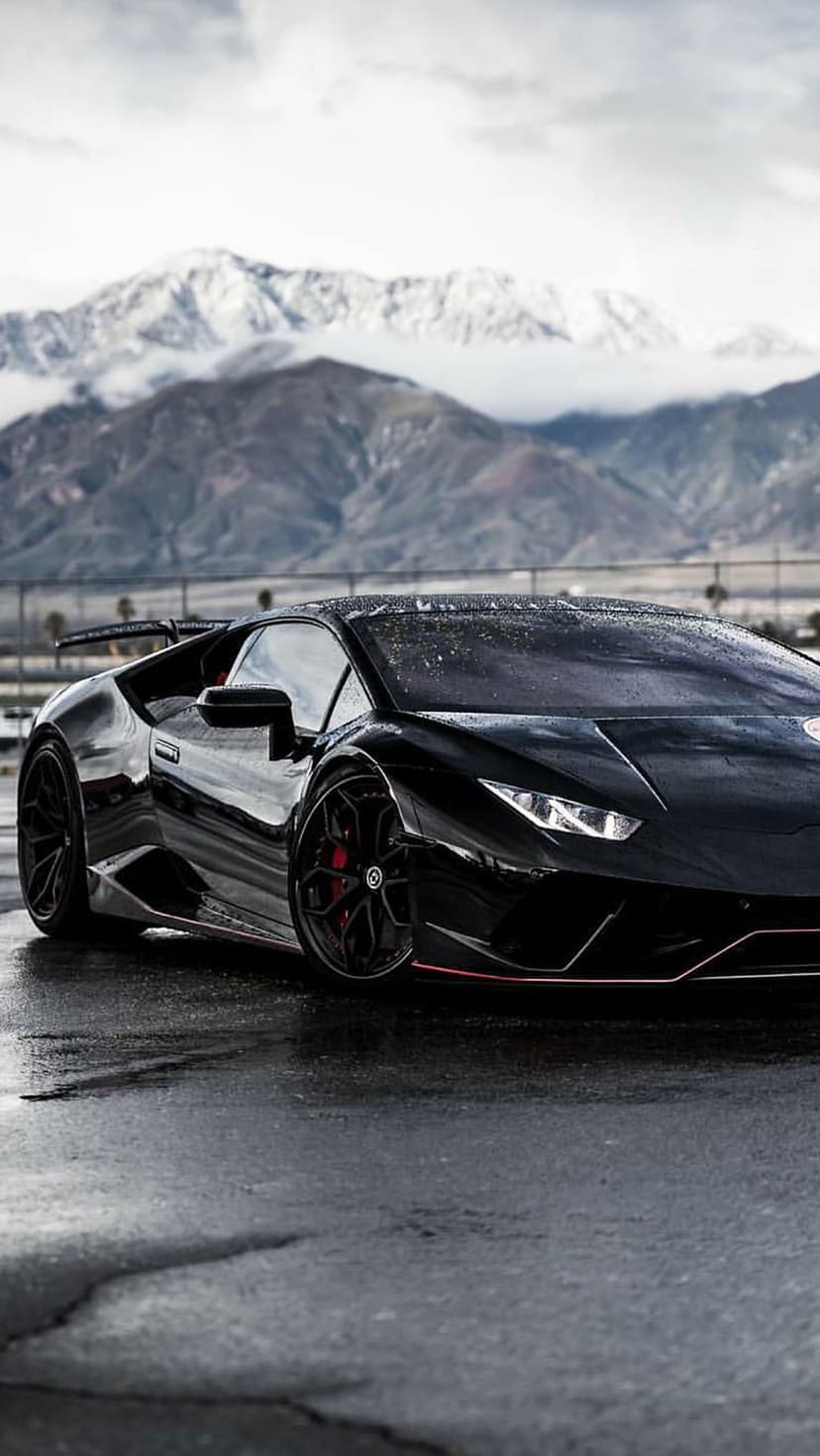 Black Lamborghini supercar on wet asphalt with red brake calipers and snow-capped mountain peaks in the distance.