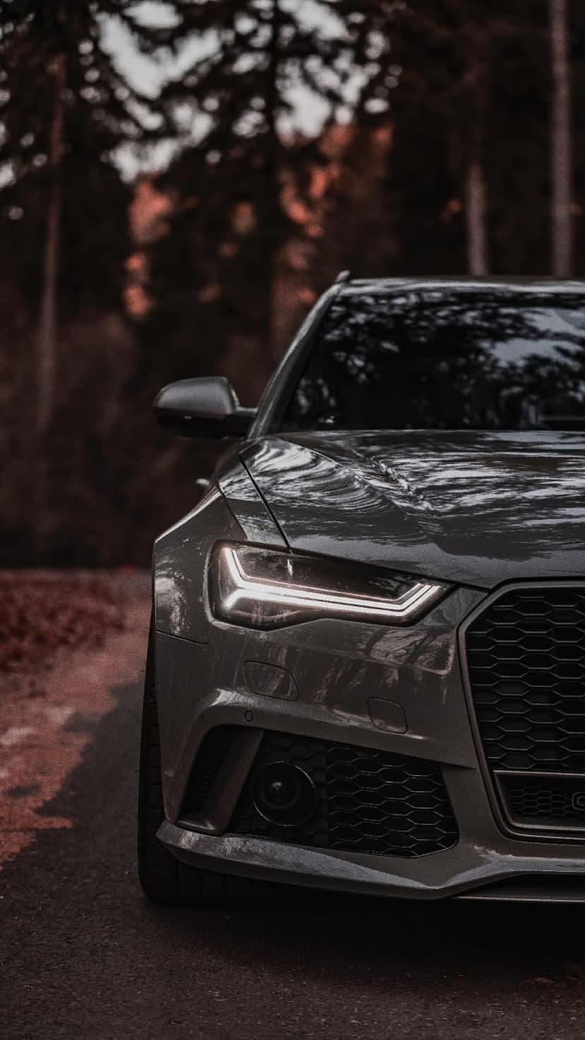 Audi RS6 Avant front grille and glowing LED headlights in a dark autumnal forest with wet surface reflections.