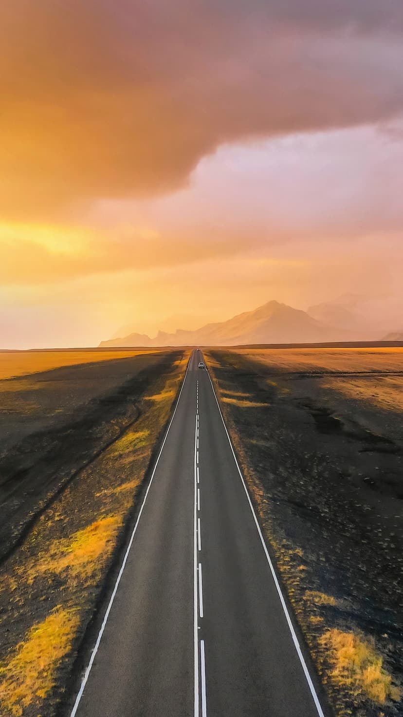 Straight asphalt road stretching through a desert toward distant mountains under a warm orange sunrise sky.