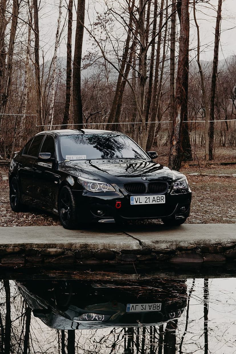 Black BMW sedan parked on a concrete platform beside a reflective lake with a dense forest in the background.