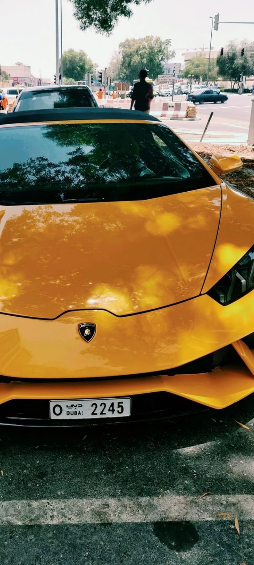 Orange Lamborghini front bumper featuring Dubai license plate 2245 and glossy paint reflecting trees and sky.