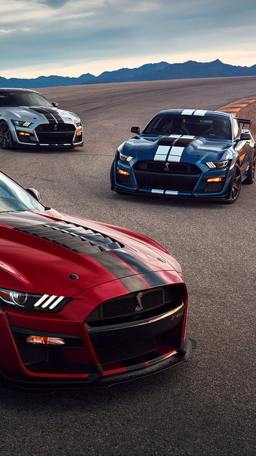 Red, blue, and silver Ford Mustang Shelby GT500 cars parked on a road with mountains and a cloudy sky.