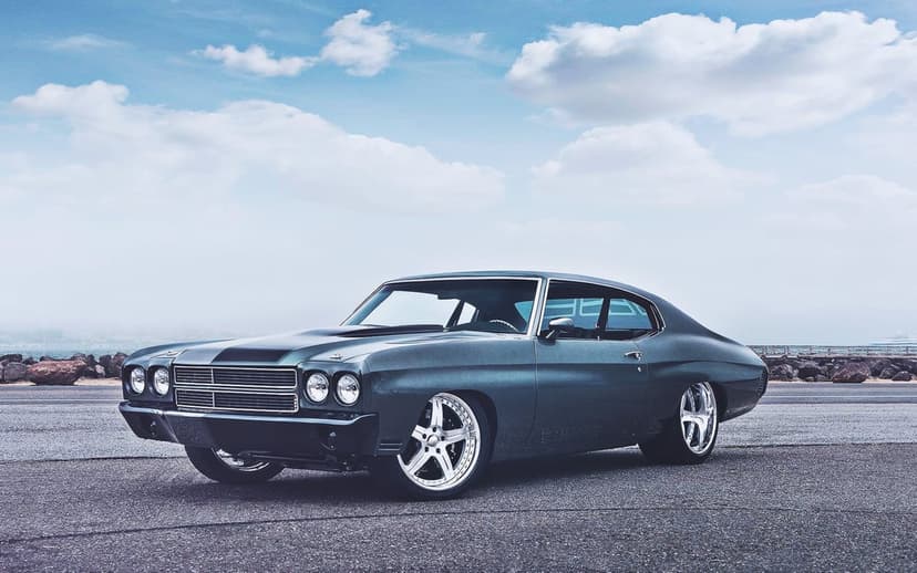 A dark grey 1970 Chevelle muscle car with chrome rims parked on asphalt under a bright cloudy sky.
