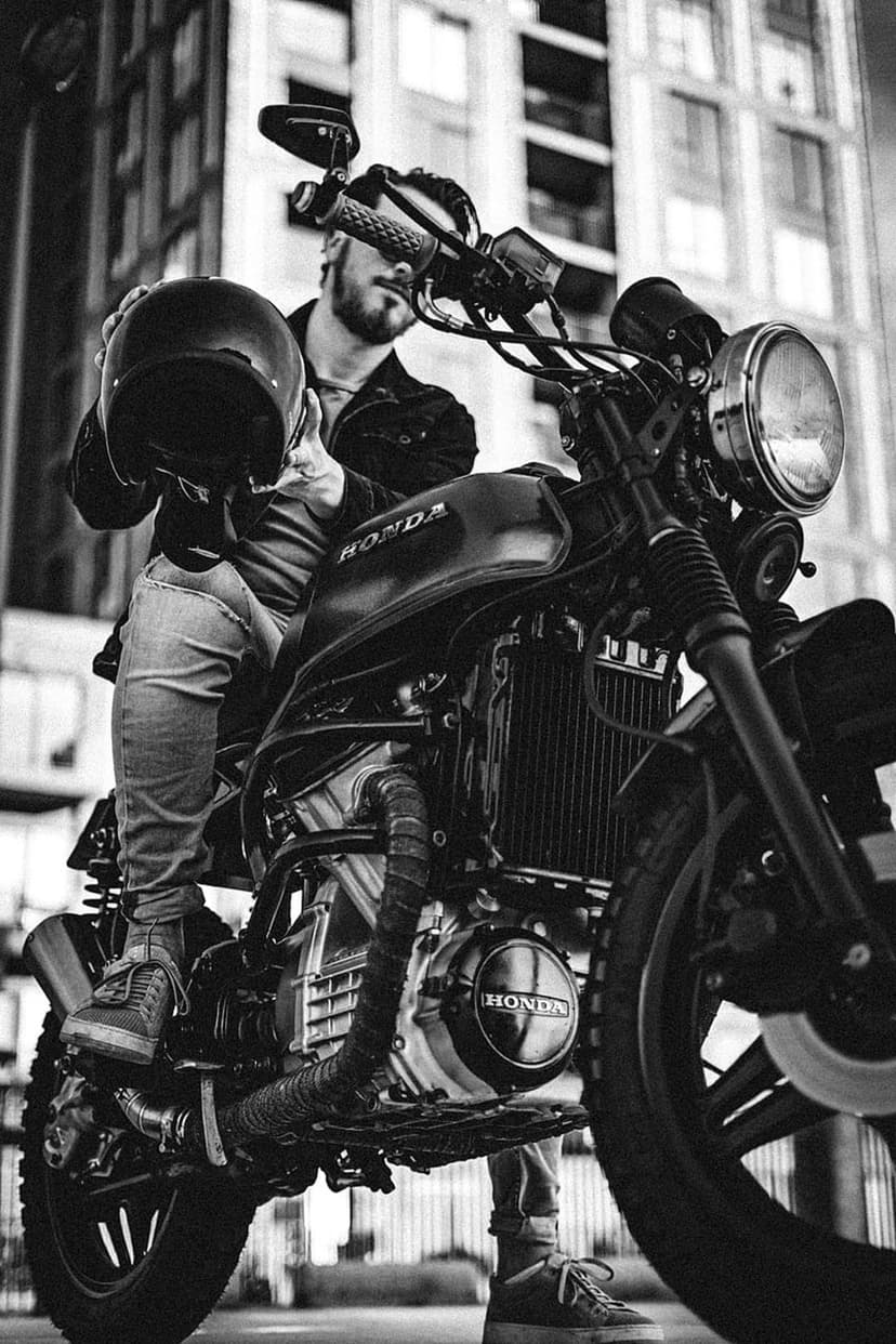 Man holding a helmet next to a vintage Honda motorcycle with urban architecture in a monochrome street setting.
