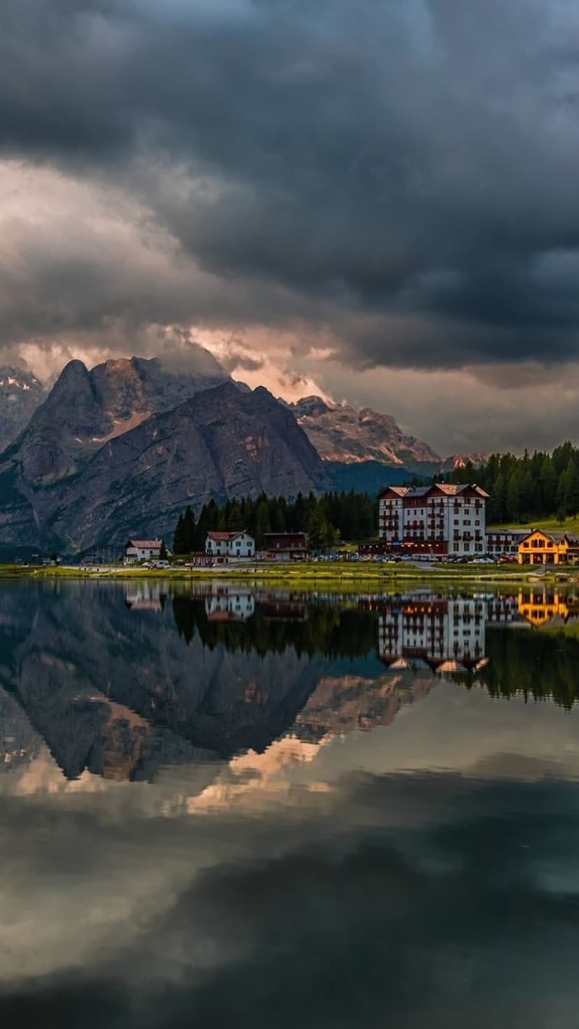 Rugged limestone mountains tower over a still lake and glowing alpine village at dusk with sharp water reflections.
