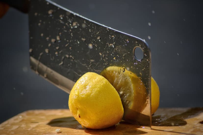 Sharp steel cleaver blade cutting through yellow lemons with water droplets splashing on a dark wooden surface.