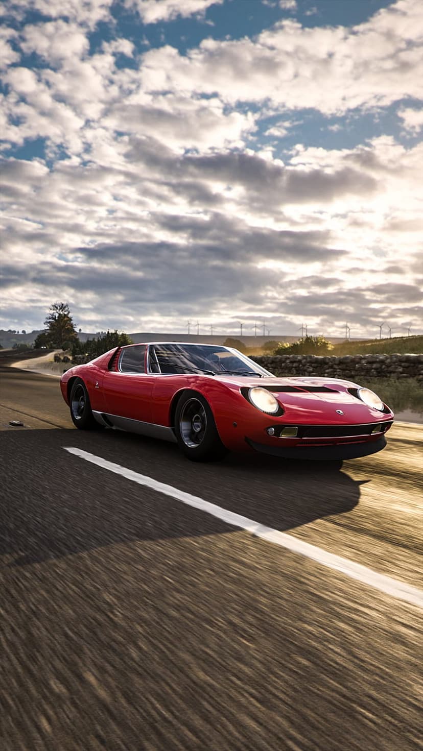 Red Lamborghini Miura driving fast on a rural road with motion blur, stone walls, and wind turbines behind it.