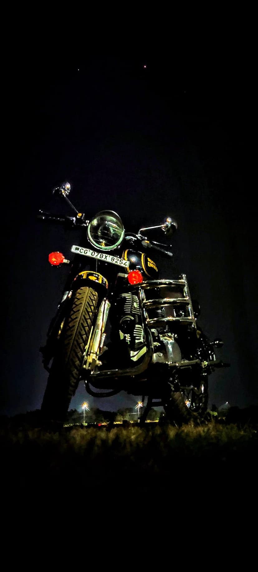 Royal Enfield motorcycle with chrome accents parked under a starry night sky with blurred urban bokeh highlights.