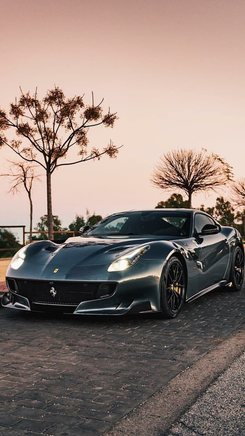 Dark grey Ferrari F12berlinetta parked on a cobblestone street beneath a warm orange sunset sky with bare trees.