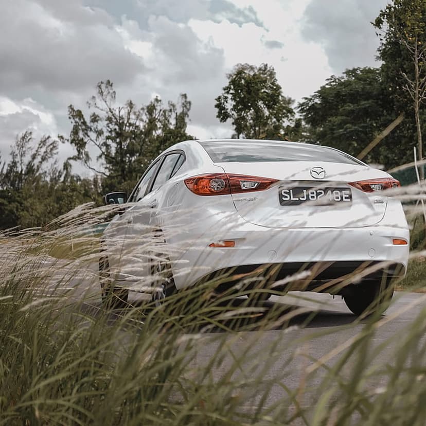 White Mazda 3 sedan rear view partially obscured by tall grass with a lush green forest and cloudy sky background.