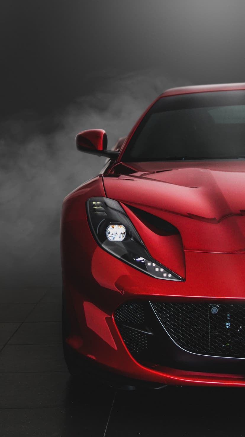 Red Ferrari headlight and front grille detail glowing in a dark, smoky setting with sharp reflections on the paint.