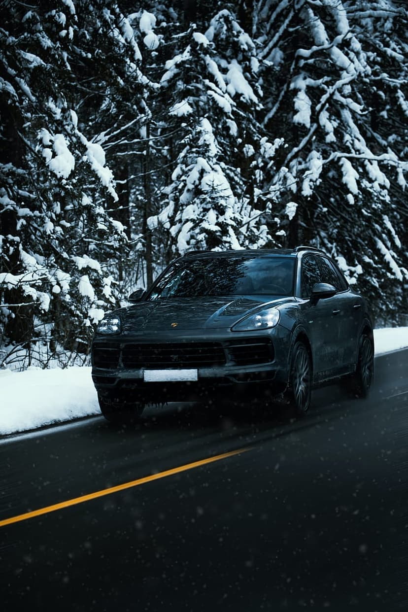 Dark luxury SUV driving on a snowy forest road with glowing yellow headlights and tall frosted pine trees.