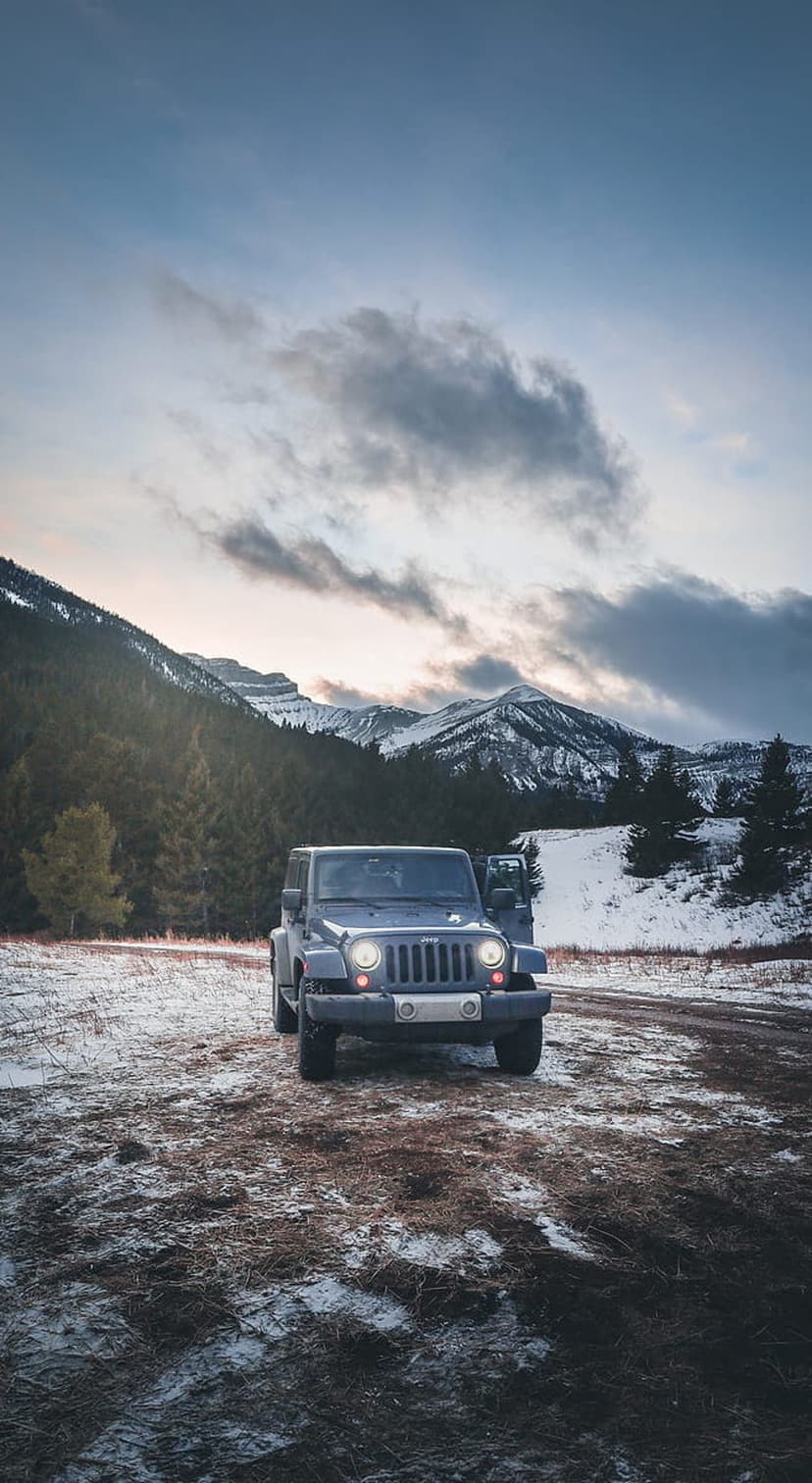 Blue Jeep Wrangler parked on a snowy dirt road with misty evergreen forests and dusk mountain peaks behind it.