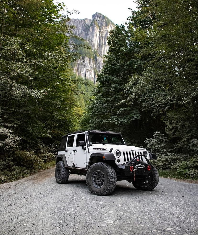 White Jeep Rubicon with off-road tires on a gravel road below a rocky mountain peak and green forest trees.