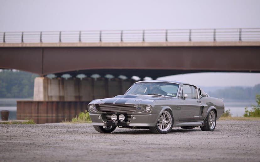 Silver 1967 Ford Mustang Eleanor with black racing stripes parked on gravel with a bridge background.