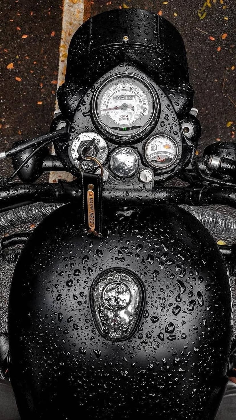 Black motorcycle dashboard with water droplets on the speedometer and a leather happiness keychain in the rain.