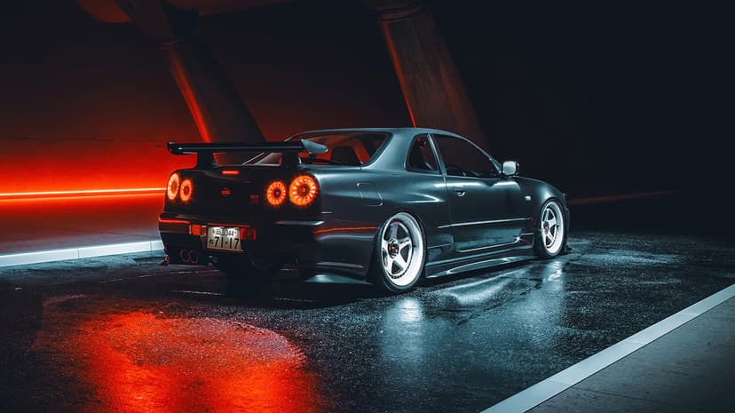 Modified dark grey Nissan Skyline GT-R R34 parked on wet asphalt under red and blue cinematic neon lights.