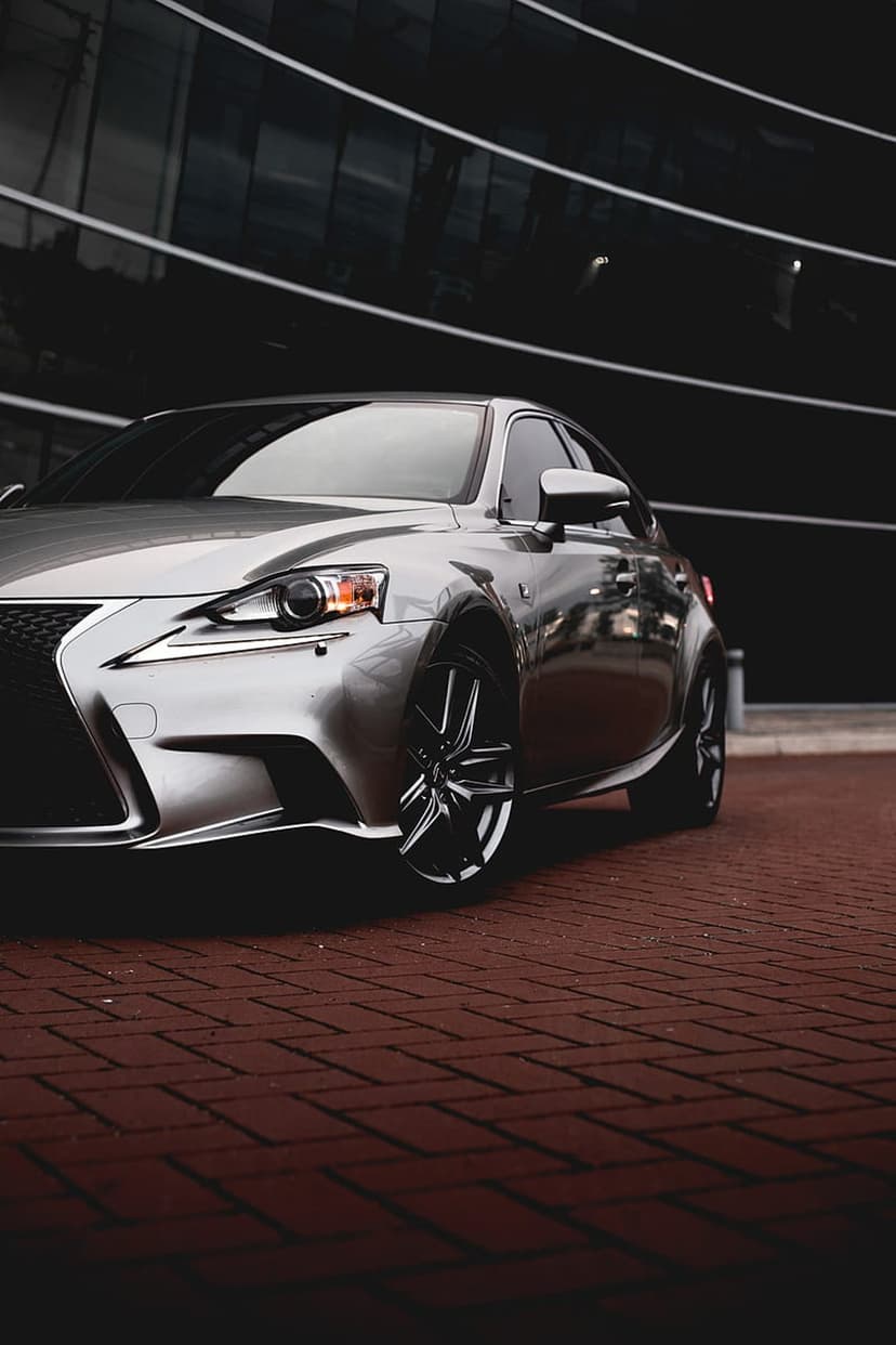 Silver Lexus IS sedan parked on a red brick patio with a modern building reflected in its metallic paint.