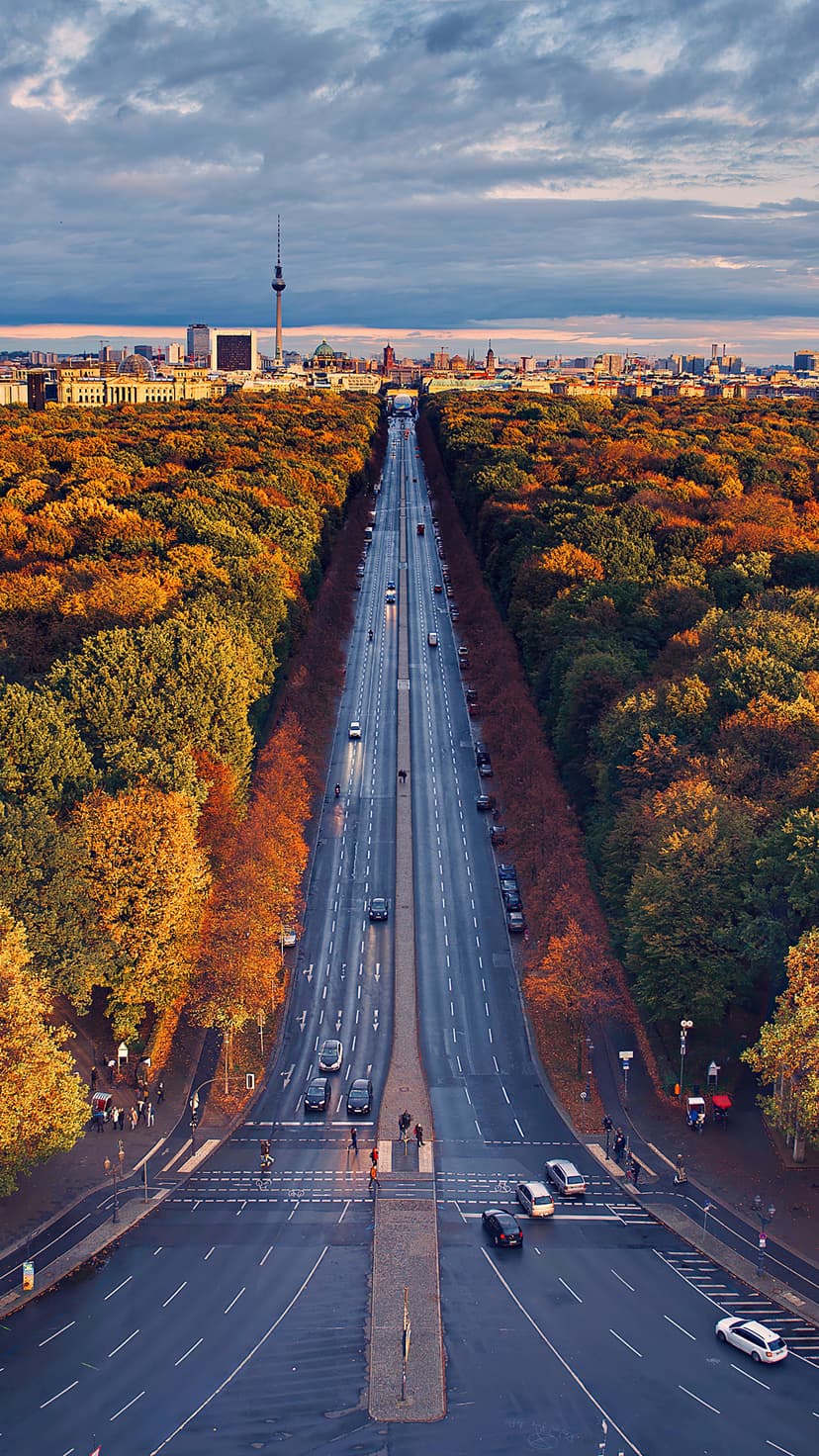 Fernsehturm Berlin stands beyond a straight boulevard bordered by dense red and gold fall foliage from above.