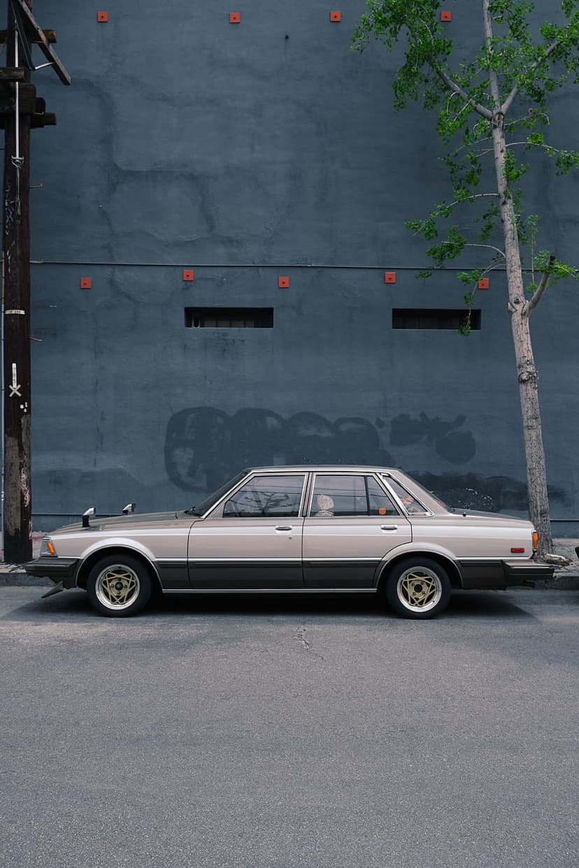 Vintage beige sedan with gold rims parked on asphalt in front of a weathered blue wall and a small green tree.