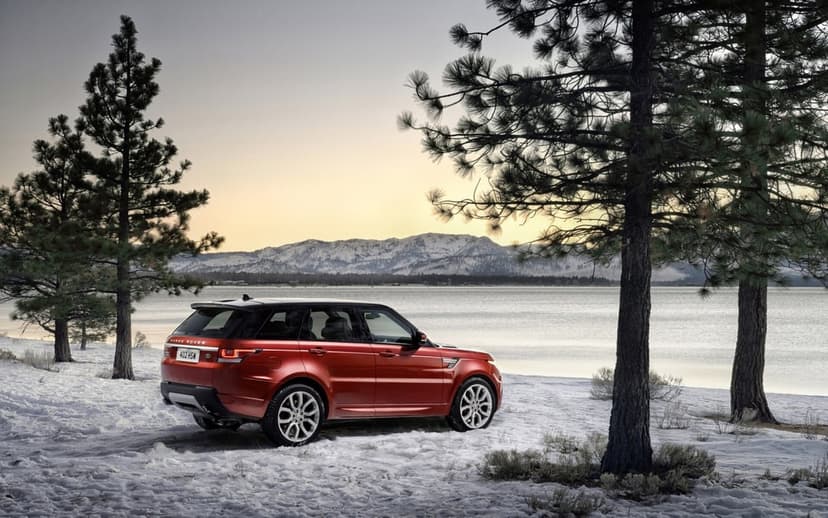 Red Range Rover Sport parked on a snowy lake shore with pine trees and snow-capped mountains at dusk.