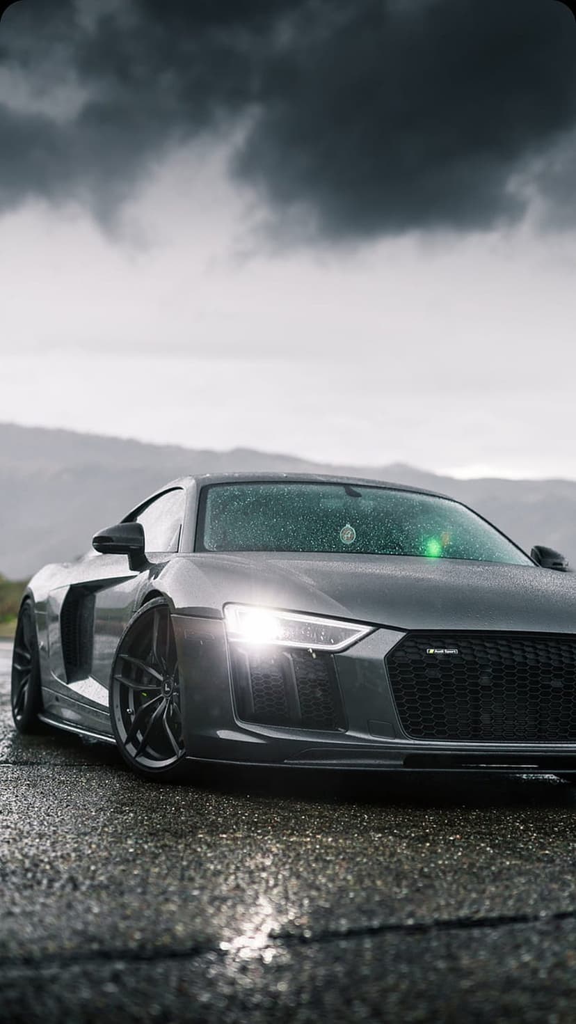 Gray Audi R8 supercar parked on a wet road at dusk with headlights on and dark storm clouds in the background.