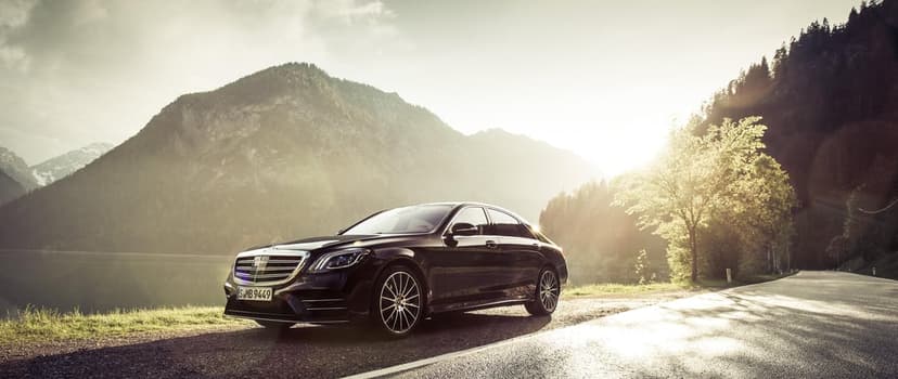 Black Mercedes-Benz S-Class luxury sedan on a mountain road beside a lake under a bright golden sunset flare.