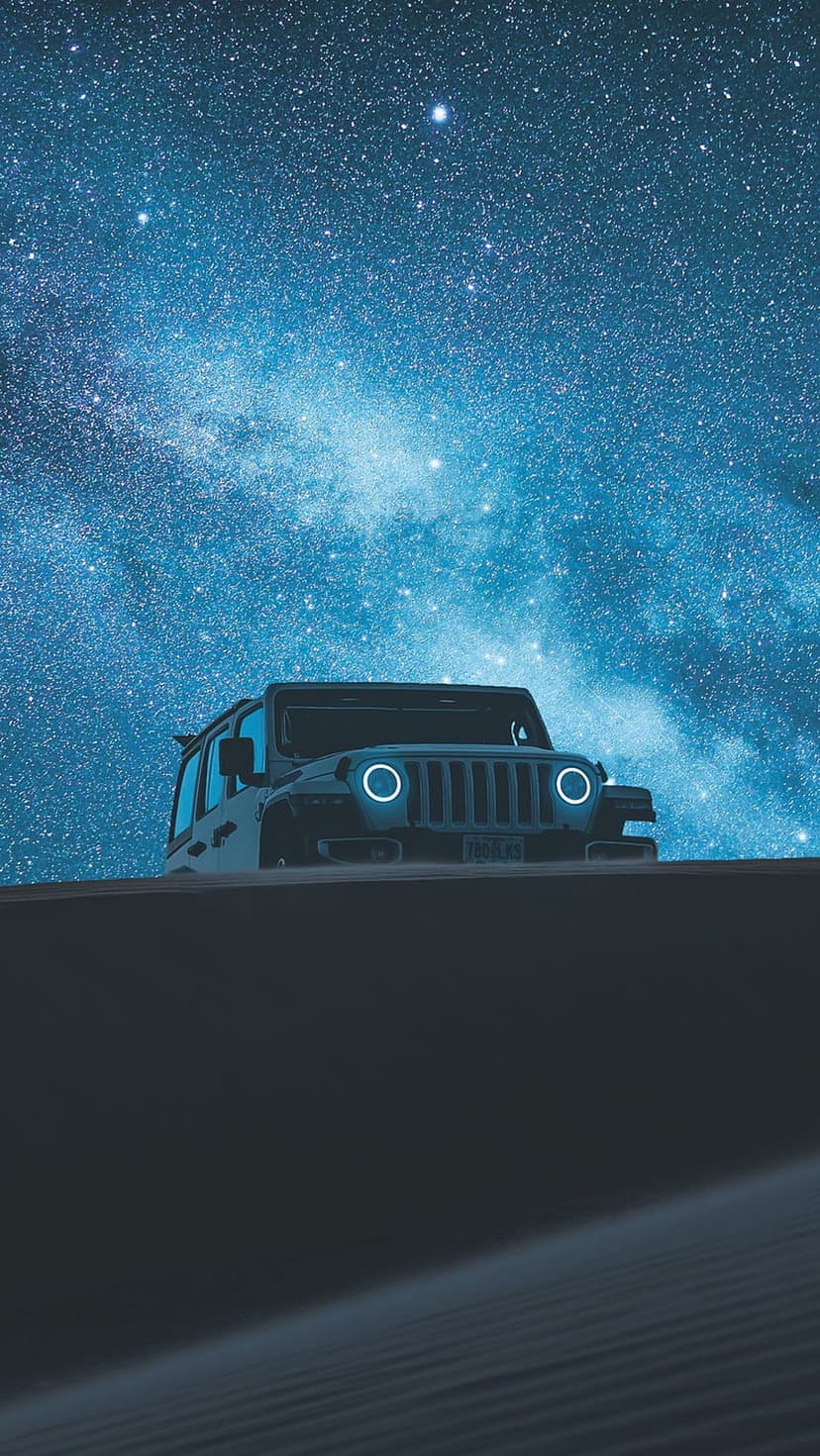Off-road Jeep Wrangler on a sand dune at night under a purple nebula and a star-filled sky with glowing headlights.