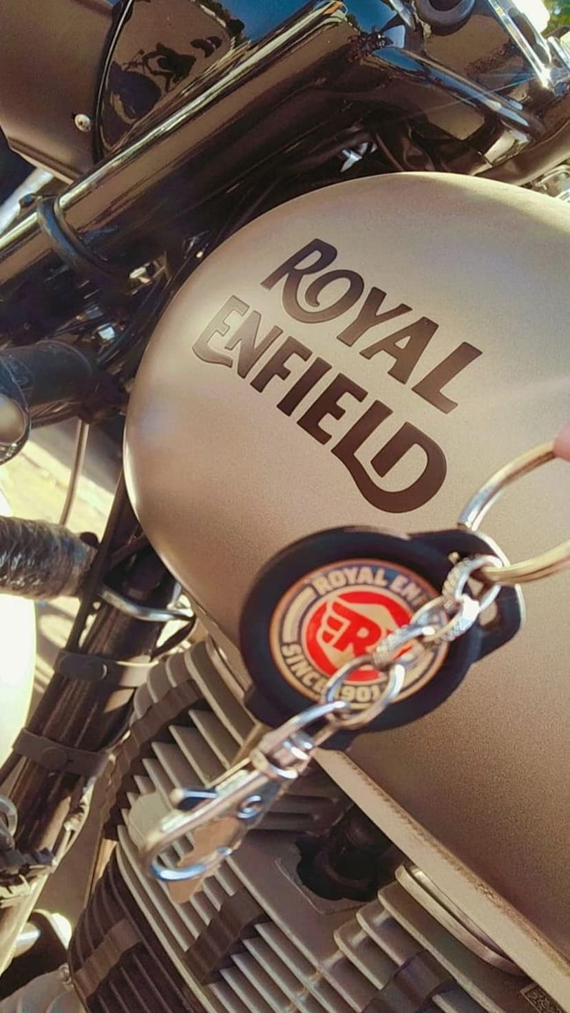 Royal Enfield silver keychain hanging from a chrome clip against a matte beige motorcycle tank in bright sunlight.