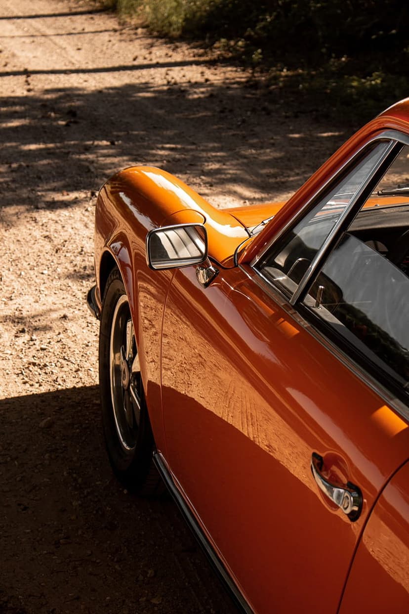 Orange vintage Porsche coupe parked on a dusty dirt road with sunlight reflecting off the polished side mirror.