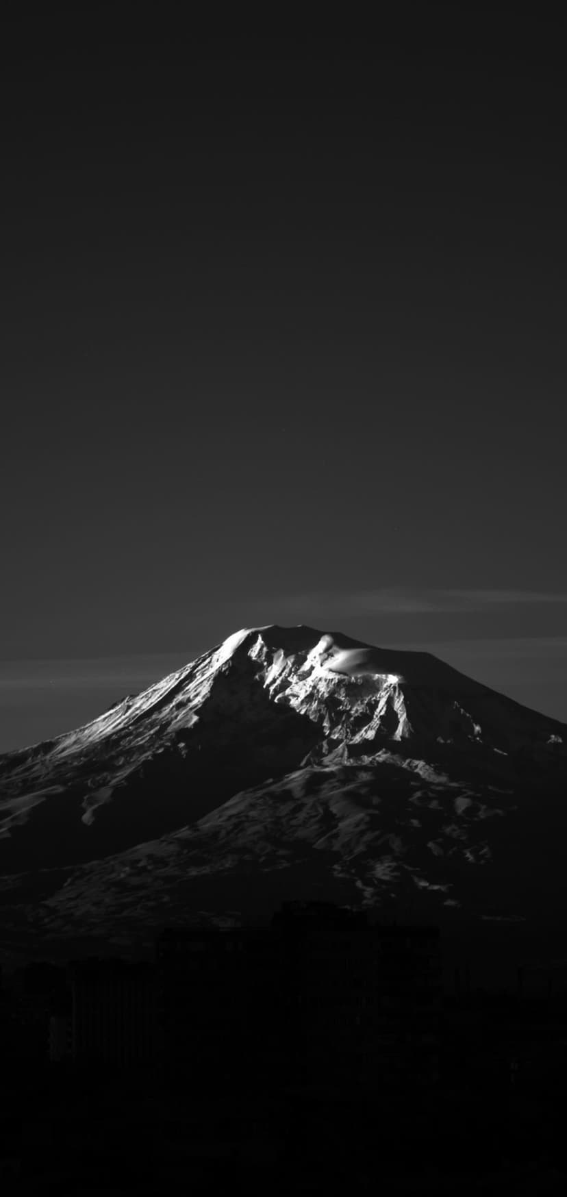 Jagged mountain peak with snow-covered slopes illuminated by a harsh beam of light against a black sky.