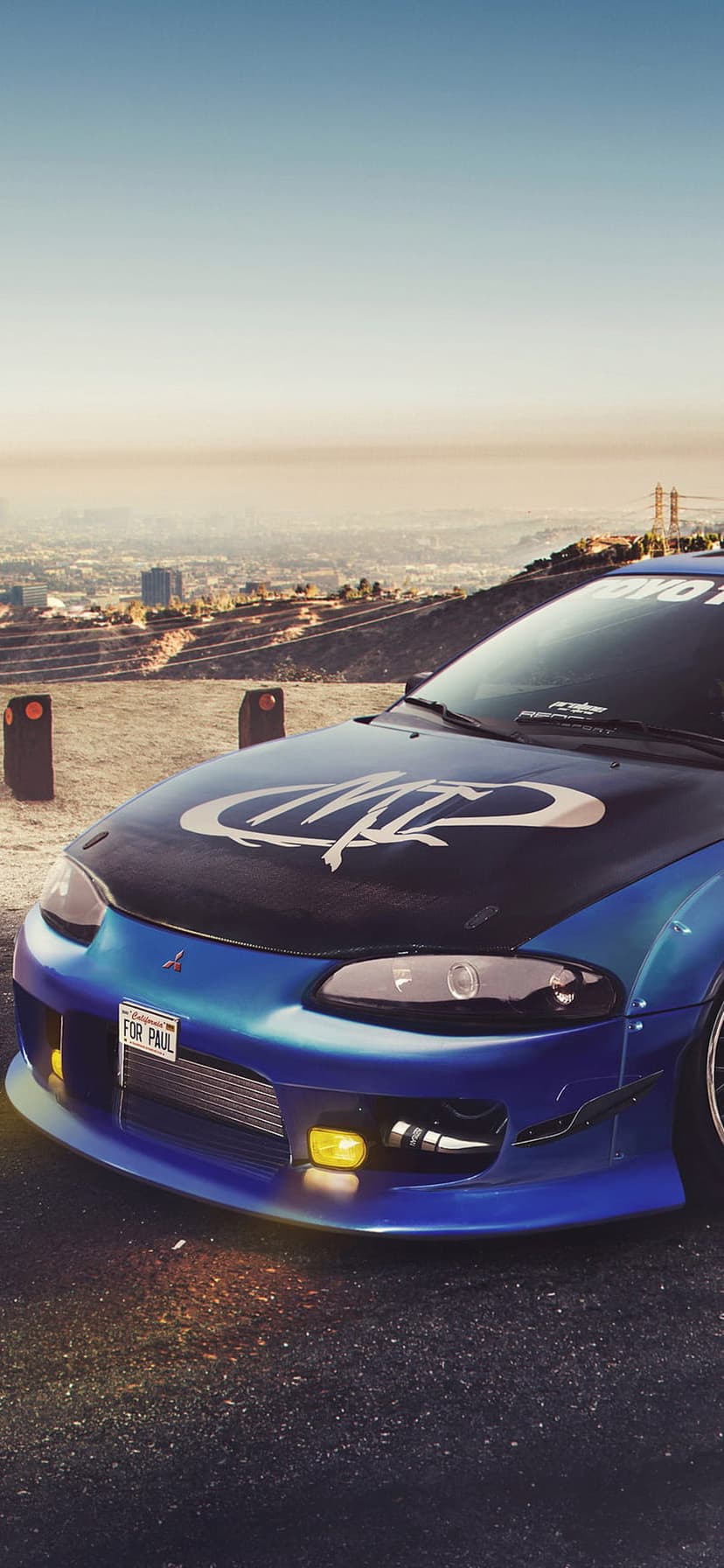 Blue Mitsubishi Eclipse with yellow fog lights parked on a hill overlooking the Los Angeles city skyline.