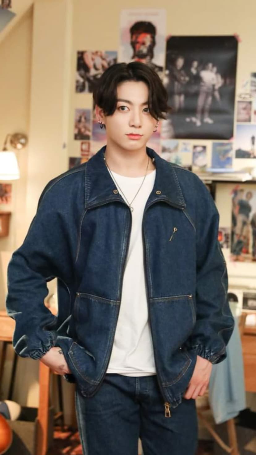 Jungkook wearing a denim jacket and jeans stands before a wall of posters in a warm, retro-style room.