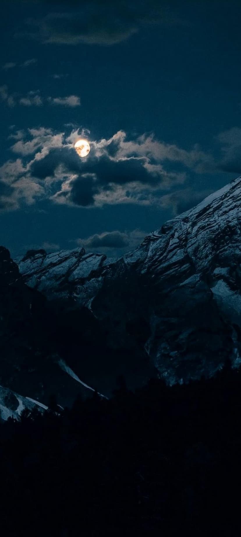 Full moon shining through dark clouds over snow-covered mountain peaks and a silhouetted forest at night.