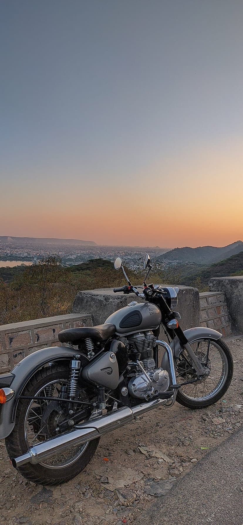 Grey Royal Enfield motorcycle on a dirt road overlooking a coastal city and lake under an orange sunset sky.