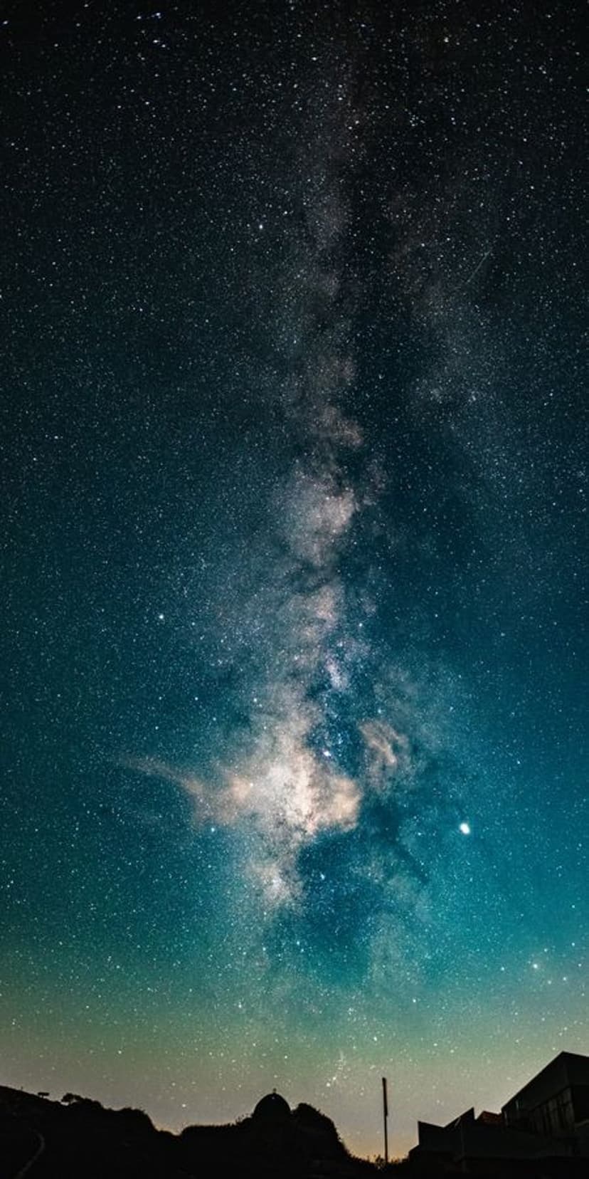 Milky Way stars stretching down to a bright turquoise horizon with dark land silhouettes in the foreground.
