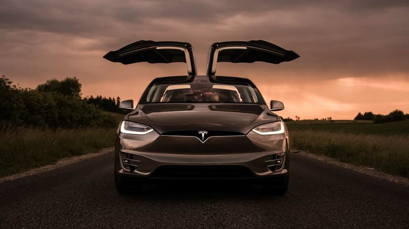 Front view of a bronze Tesla Model X with falcon wing doors open on a road during a cloudy orange sunset.