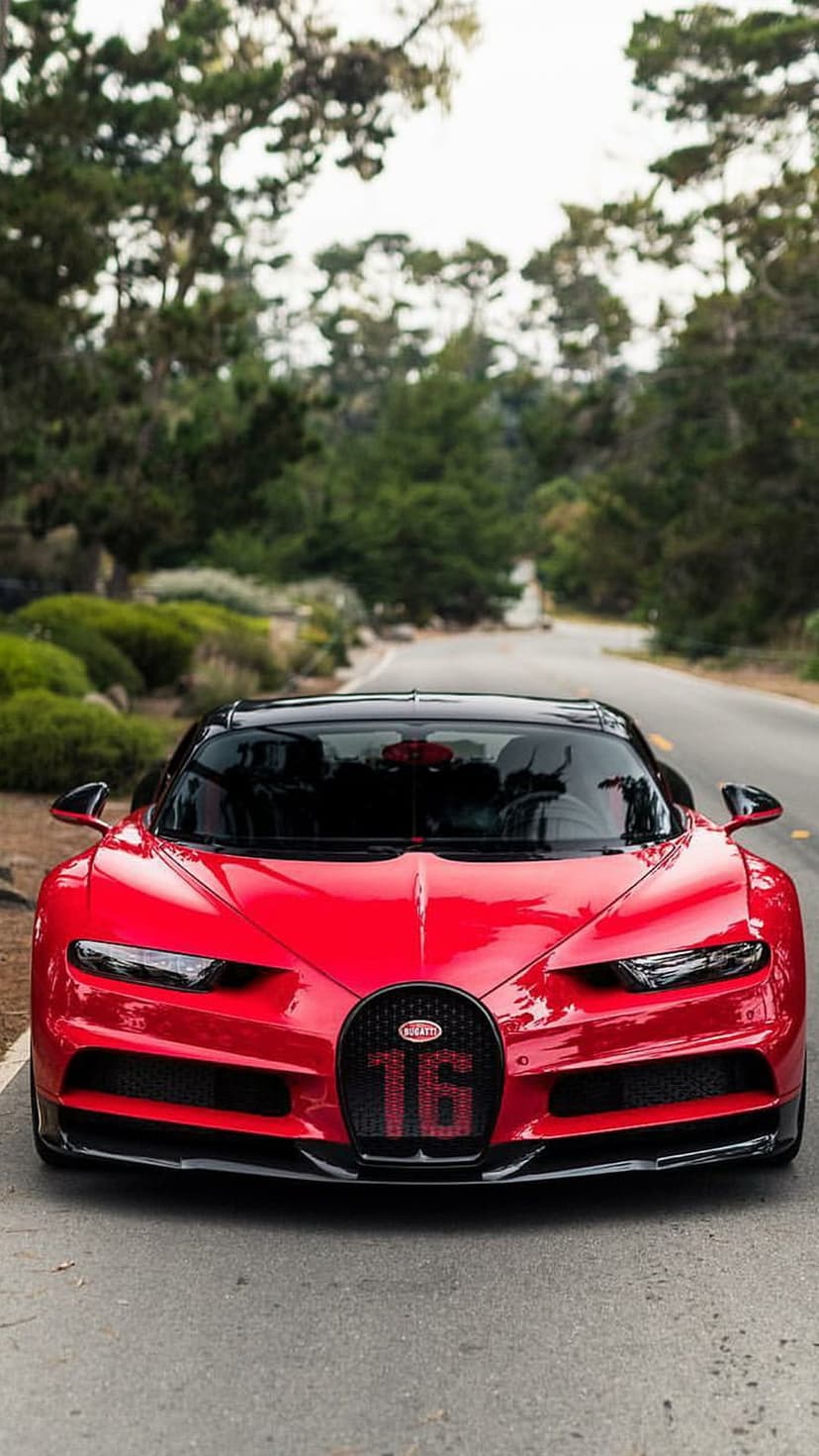 Red Bugatti Chiron supercar with a 16-emblem grille parked on a tree-lined road in a vertical composition.