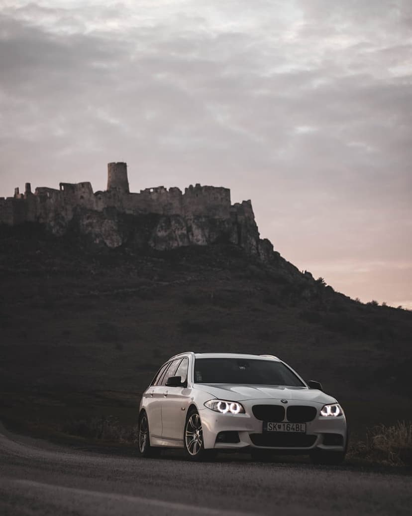 White BMW station wagon with glowing headlights faces ancient stone castle ruins under a dark overcast sky.