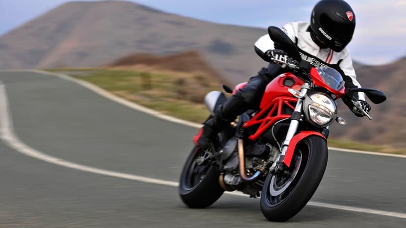 Red Ducati Monster motorcycle leaning into a mountain road turn with a rider in white and black leather gear.