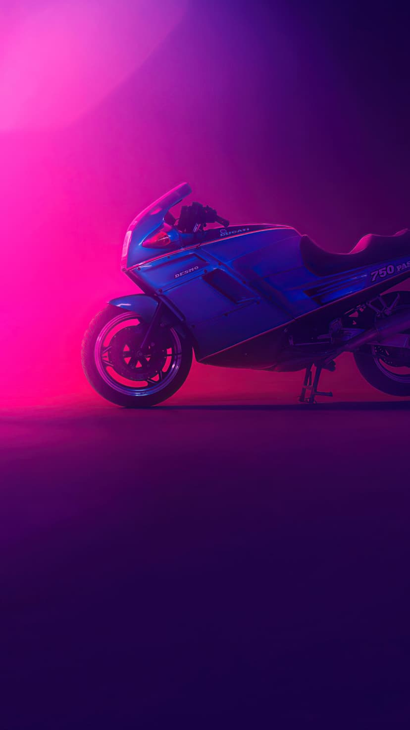 Blue vintage sportbike silhouetted against neon pink and purple smoke with glowing highlights on the chrome.