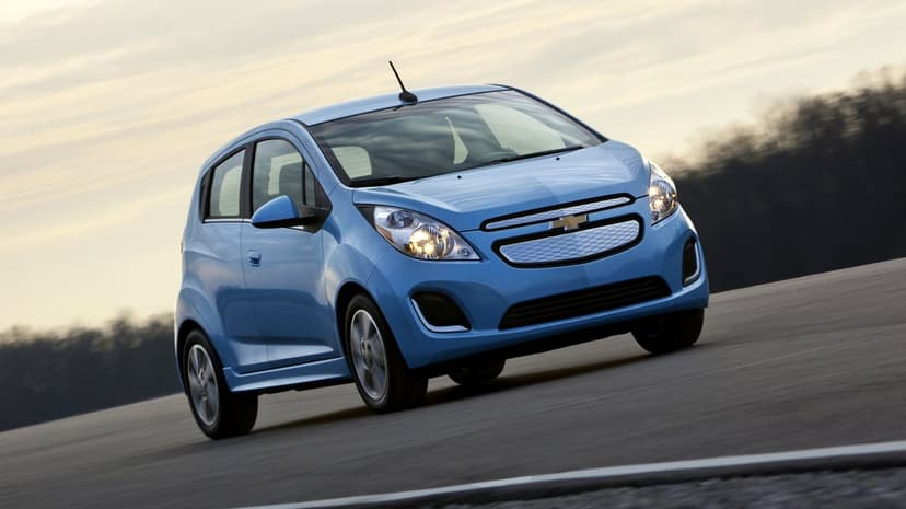 Light blue Chevrolet Spark EV driving fast on a gray asphalt road with blurred trees under a pale hazy sky.