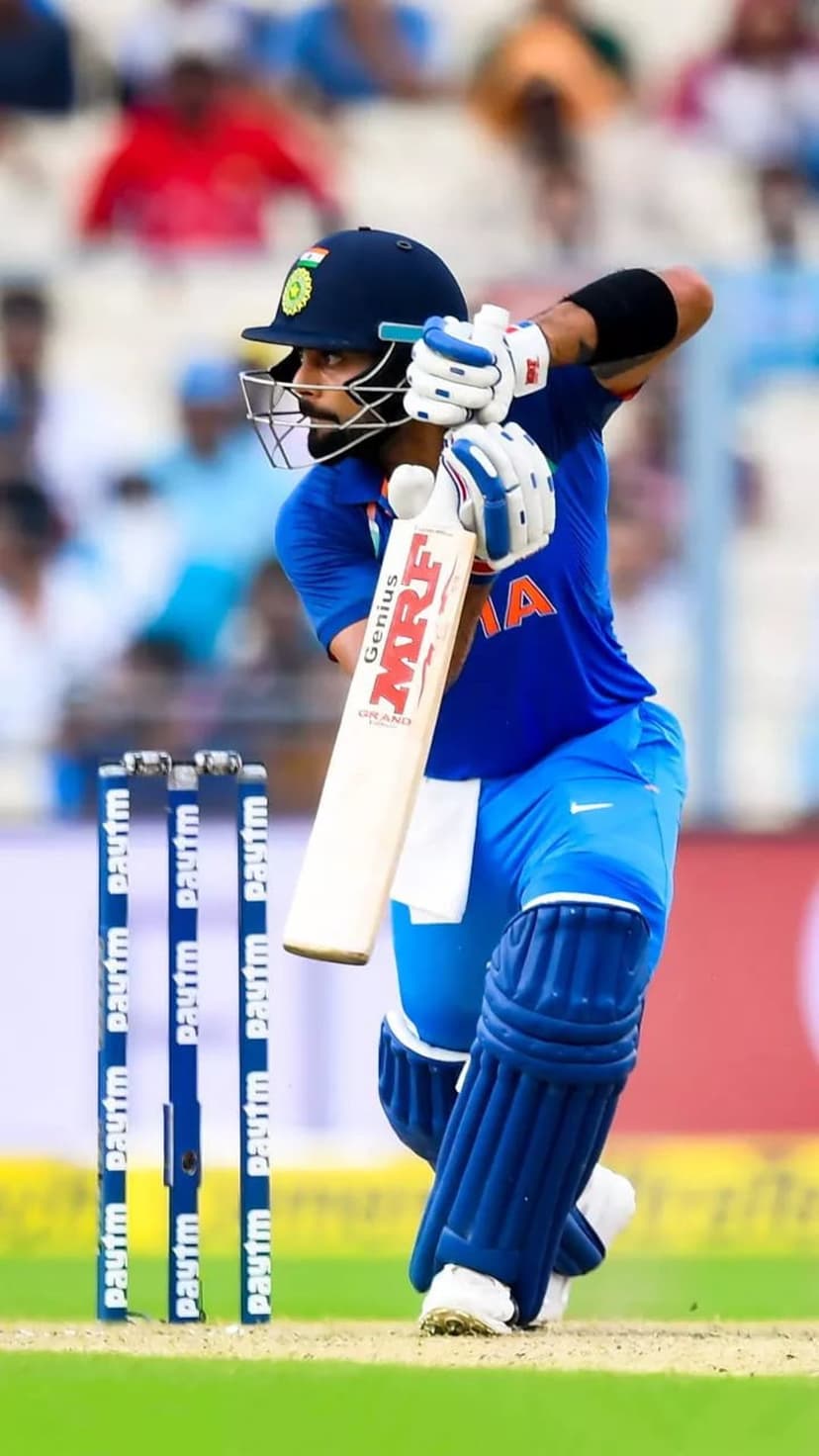 Virat Kohli in a blue cricket jersey holds a bat in a powerful stance on a green grass field near wickets.