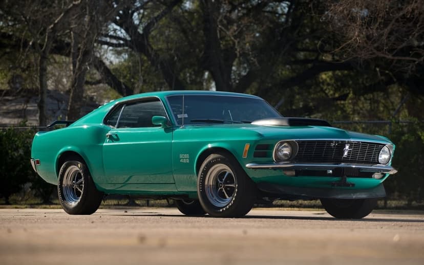Teal 1970 Ford Mustang Boss 429 with a hood scoop parked on asphalt with blurred trees in the background.
