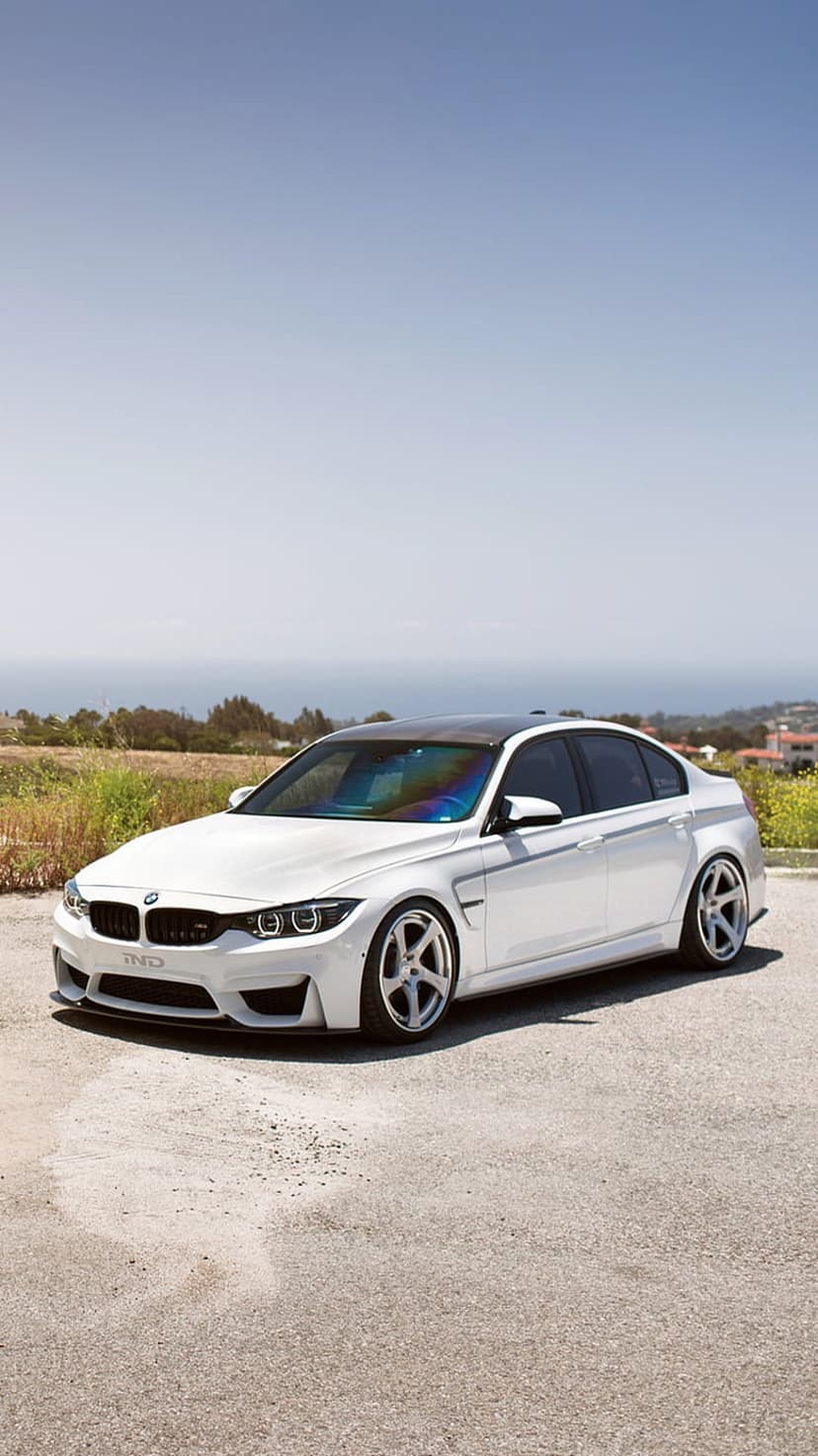 White BMW M4 Coupe with black wheels parked on a sunny coastal road overlooking the ocean and green hills.