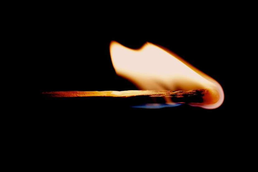 Matchstick head igniting with bright orange fire, blue heat at the base, and glowing embers on a dark backdrop.